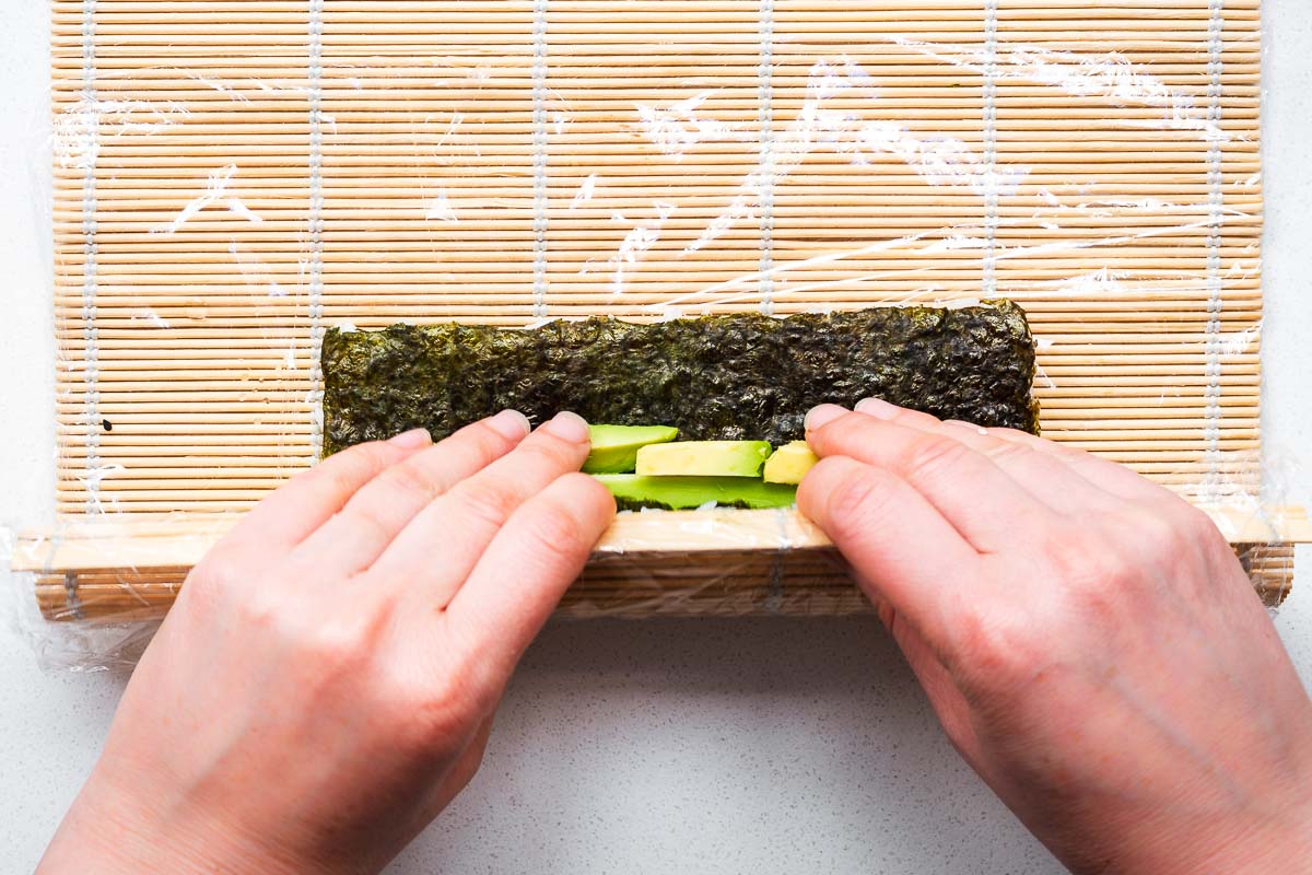 A hand rolling inside out sushi rolls on a plastic-covered bamboo sushi mat.