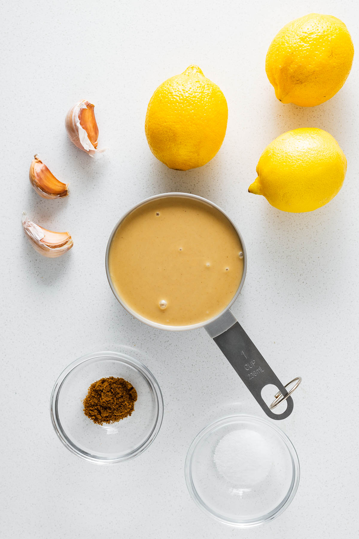 The simple ingredients for easy lemon tahini sauce viewed from above. This includes fresh lemons, tahini, garlic cloves, ground cumin and kosher salt.