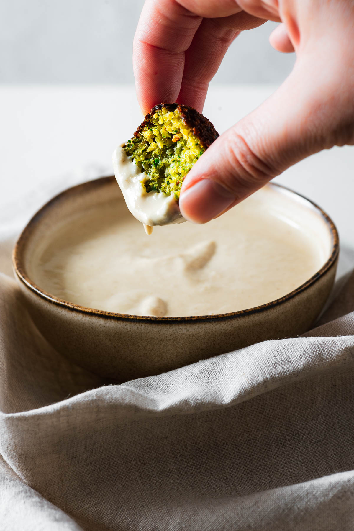A close-up of a hand dipping a fresh Middle Eastern falafel into a small bowl of tahini sauce.