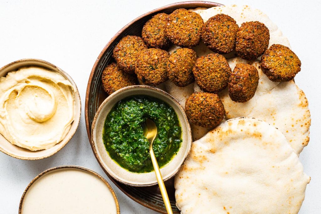 Falafel patties on a platter with hummus, zhoug, tahini sauce and white pita bread.