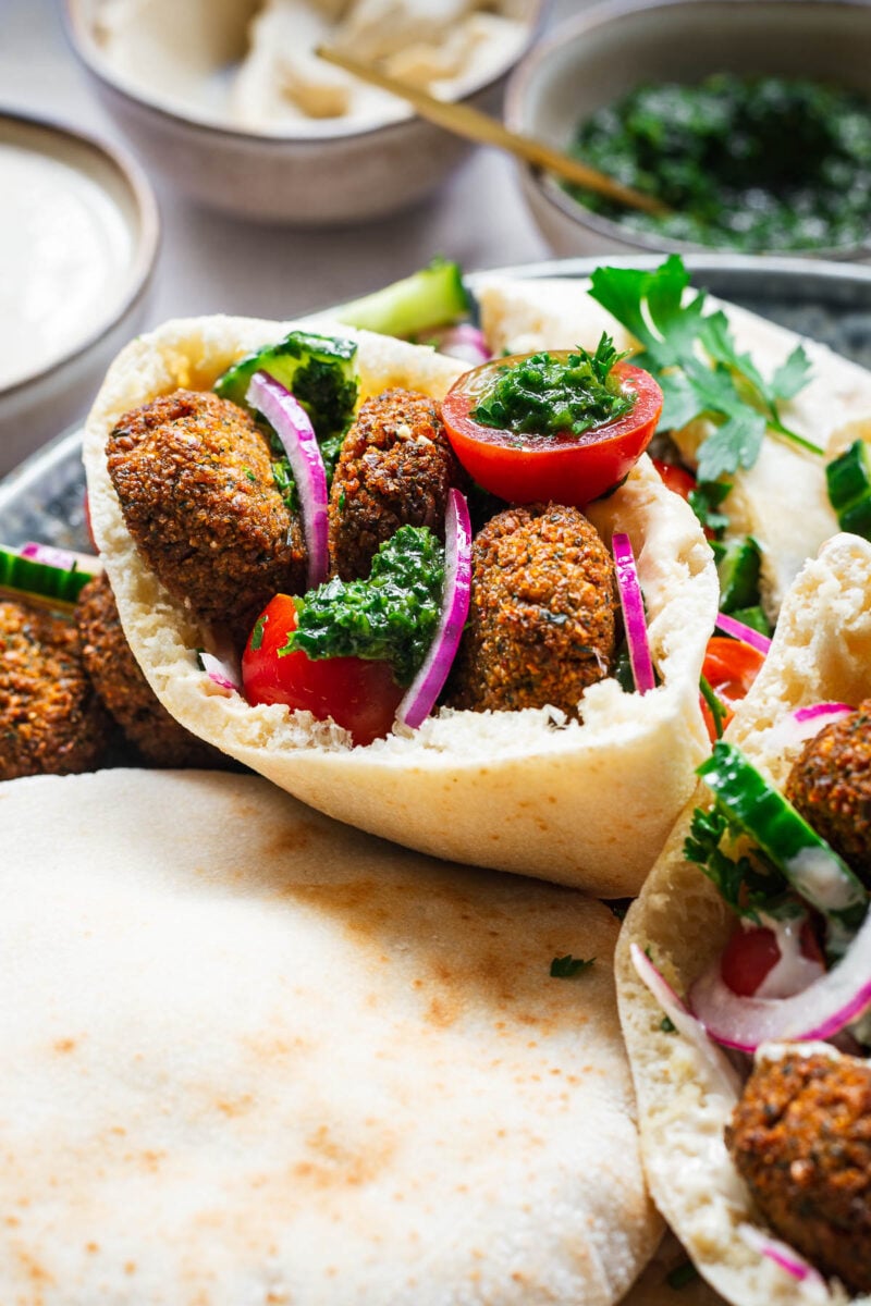 Close-up of a falafel pita sandwich with red onion, cherry tomato, cucumber, parsley, tahini sauce and zhoug hot sauce.