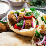 Close-up of a falafel pita sandwich with hummus, chopped vegetables and herbs, tahini sauce and zhoug hot sauce.