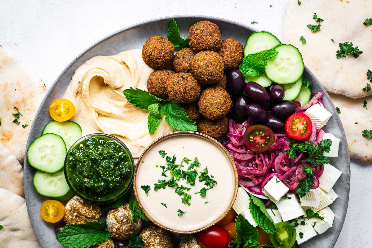 A falafel platter with falafel balls, hummus, tahini sauce, sliced cucumber, tomatoes, feta cheese, labneh balls, olives and pita bread.