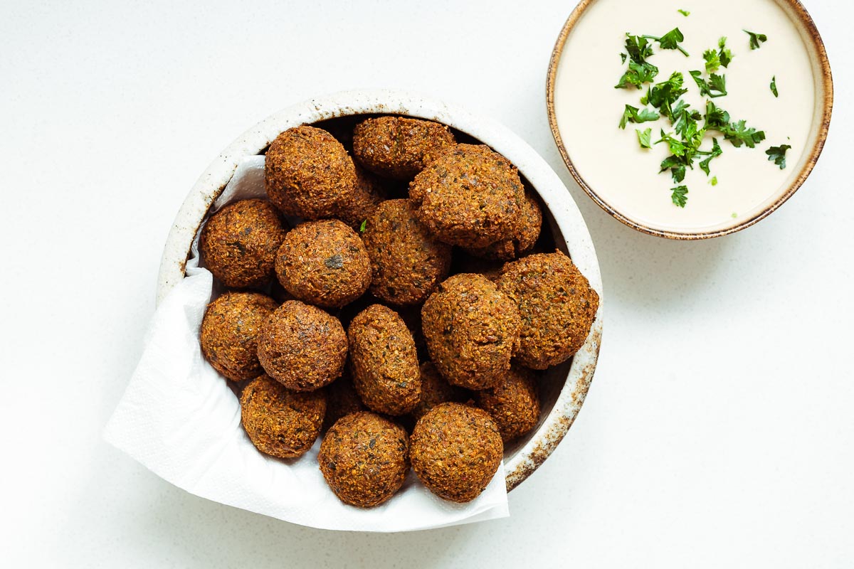 A ceramic bowl with crispy falafel balls and falafel patties next to a small bowl of falafel tahini sauce viewed from above.