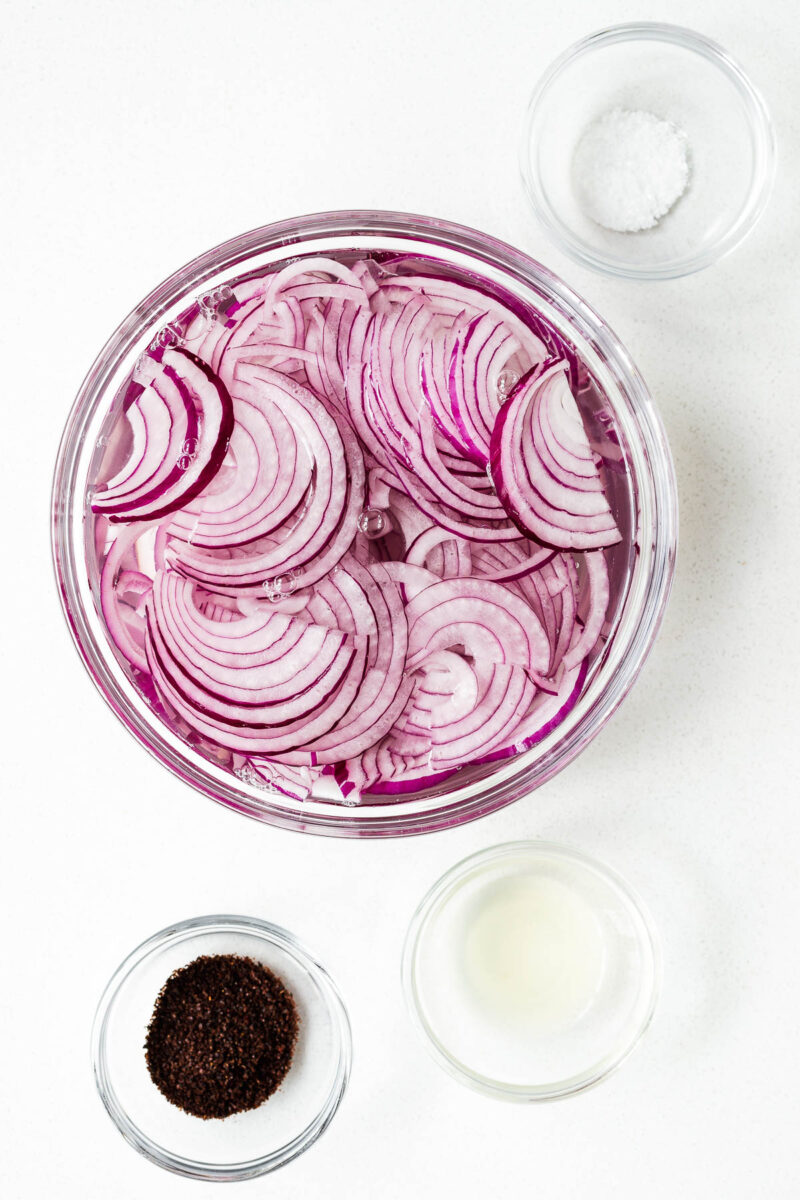 Thinly sliced red onions in a bowl with cold water with small bowls of sumac, lemon juice and salt.