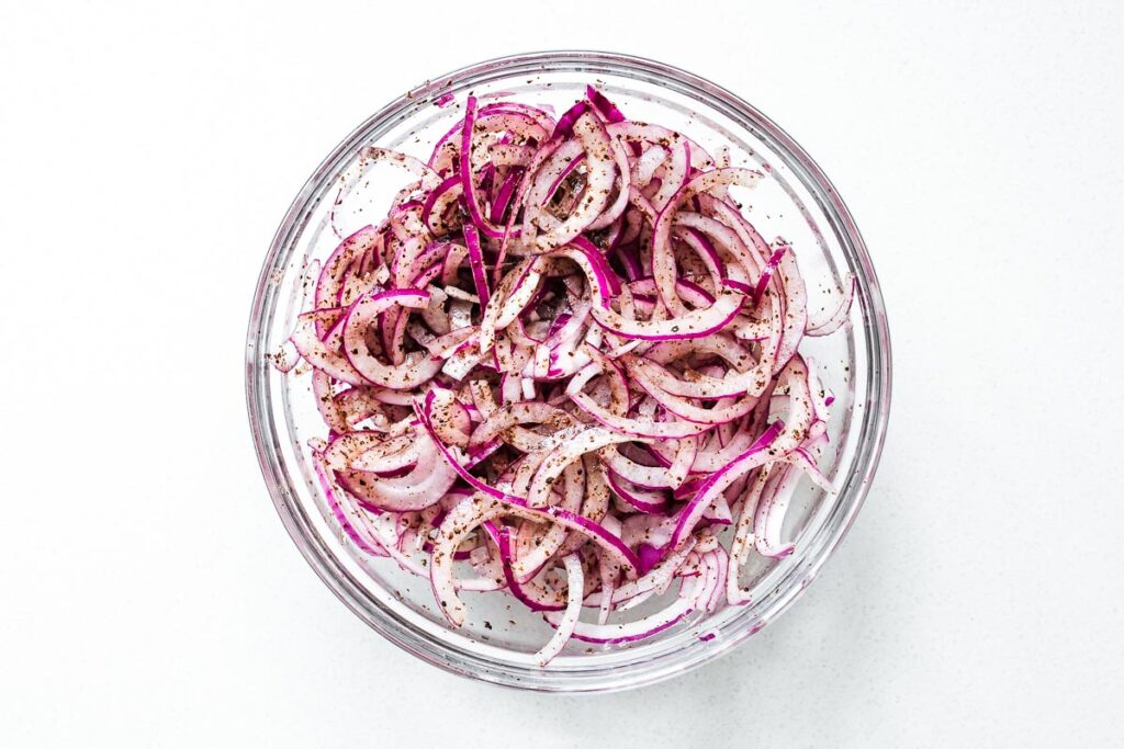 Thinly sliced red onions with sumac and lemon juice in a large mixing bowl.