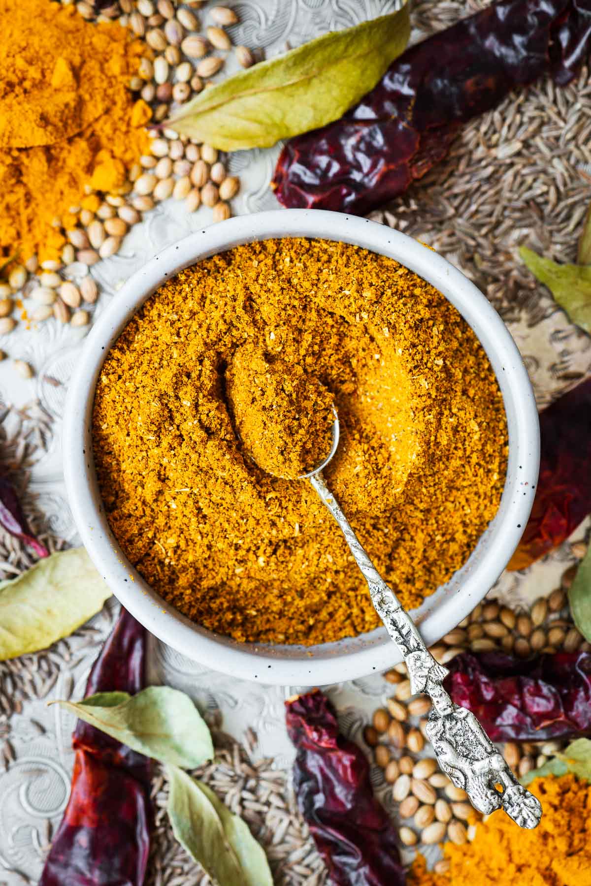 Homemade curry powder in a small bowl with a serving spoon surrounded by ingredients such as turmeric, coriander, cumin, chillies, black pepper and dried curry leaf.