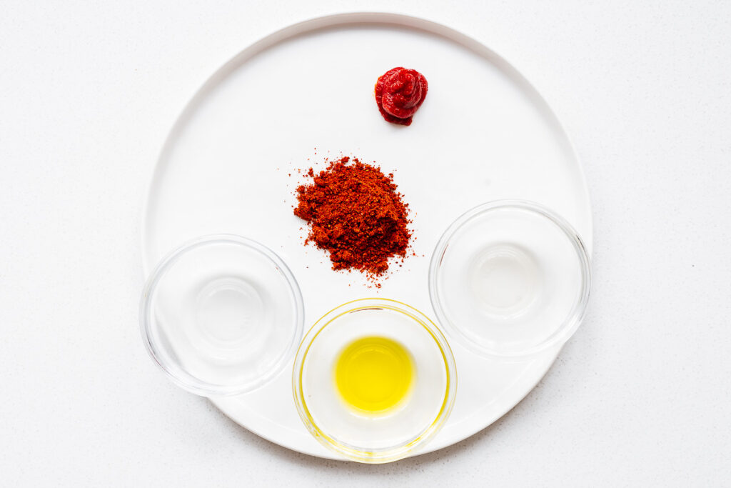 Ingredients required to make harissa paste from harissa powder also include water, vinegar (or lemon juice), olive oil and tomato paste. The ingredients are arranged on a white plate viewed from above.