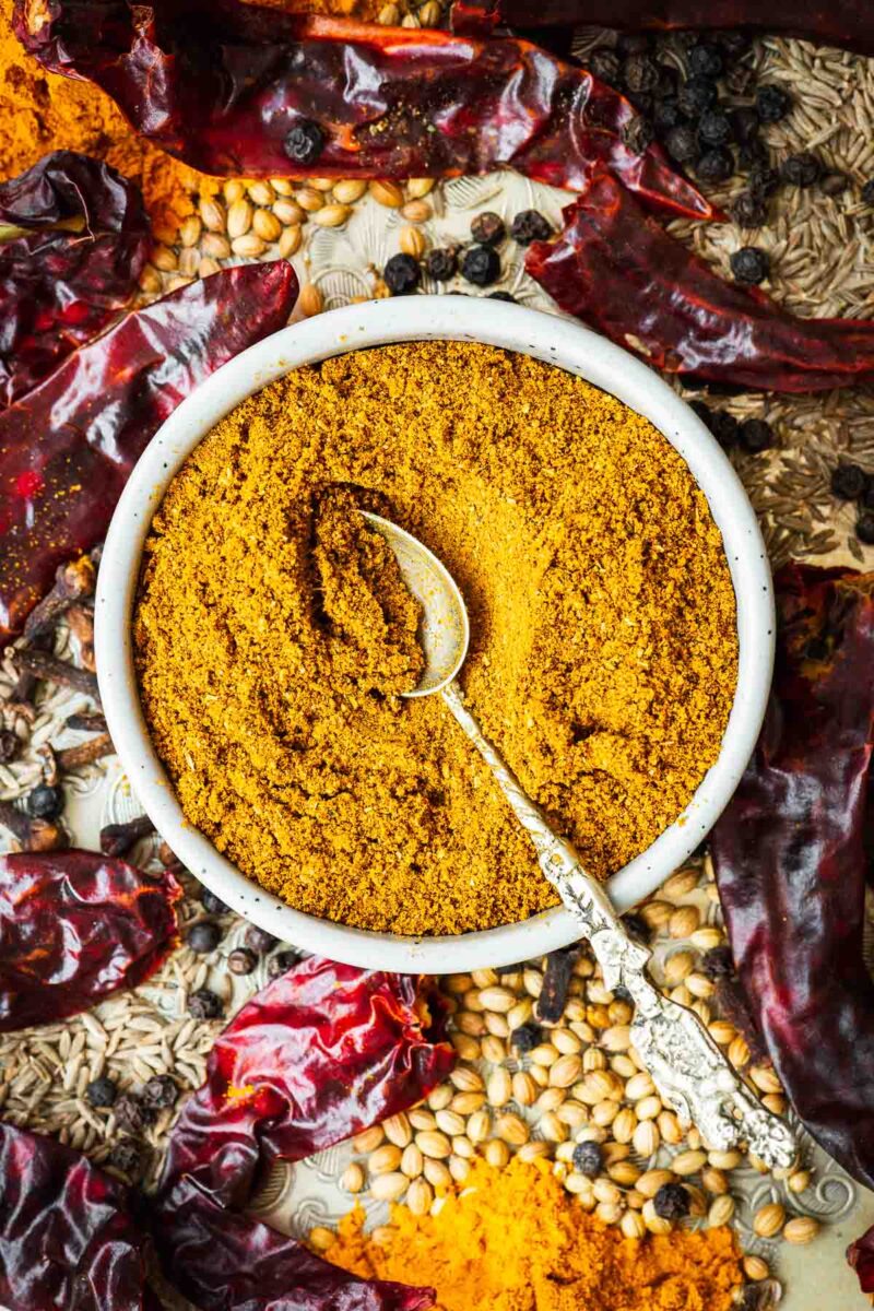 Madras curry powder in a small ceramic bowl with an ornate teaspoon surrounded by curry powder ingredients.