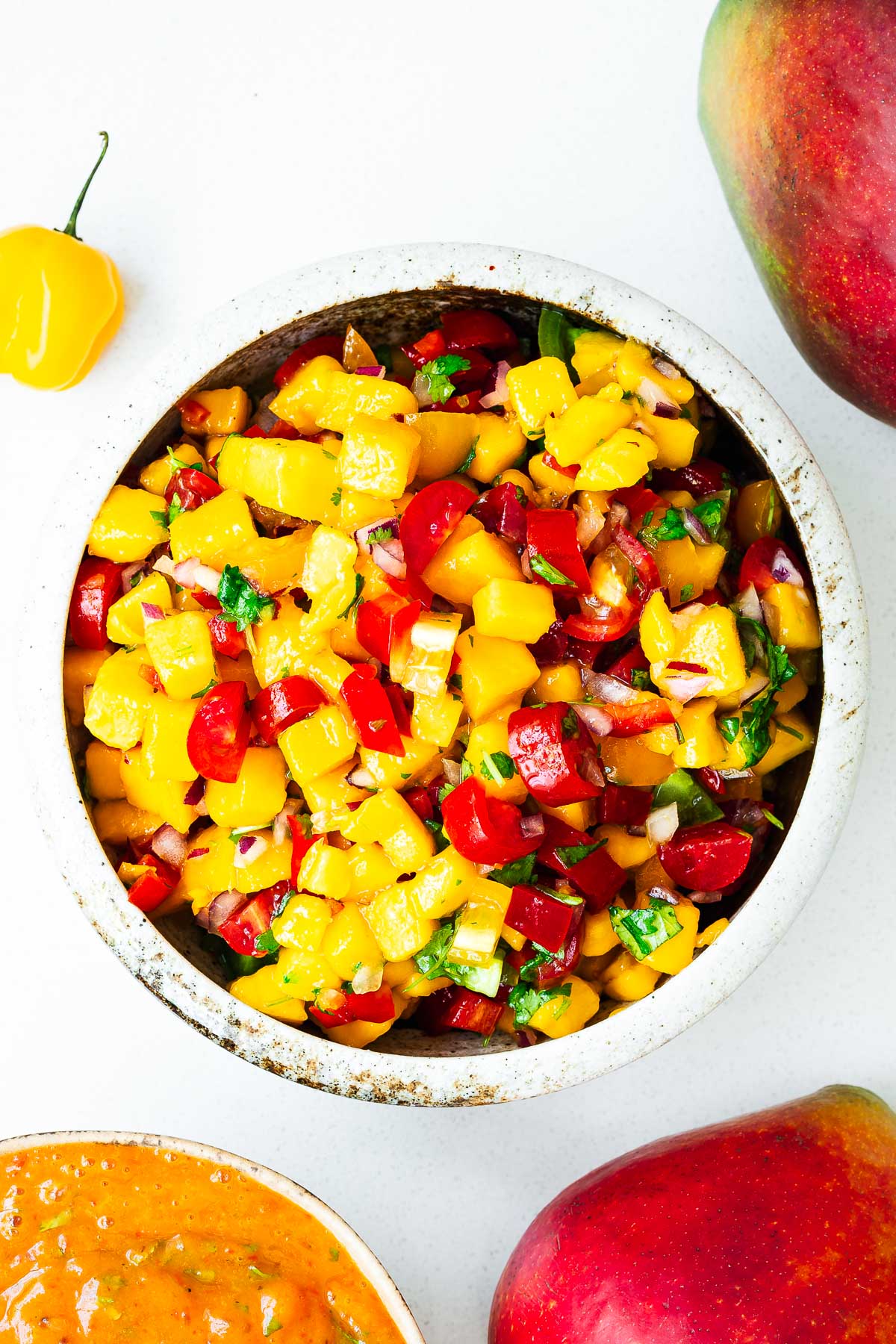 A bowl with fresh mango habanero salsa alongside a bowl of roasted mango habanero salsa surrounded by fresh mangoes and habanero peppers.