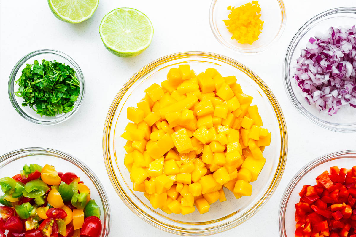 All the prepared ingredients for a fresh mango habanero salsa including diced onion, mango, tomato, red pepper, cilantro, habanero and fresh limes.