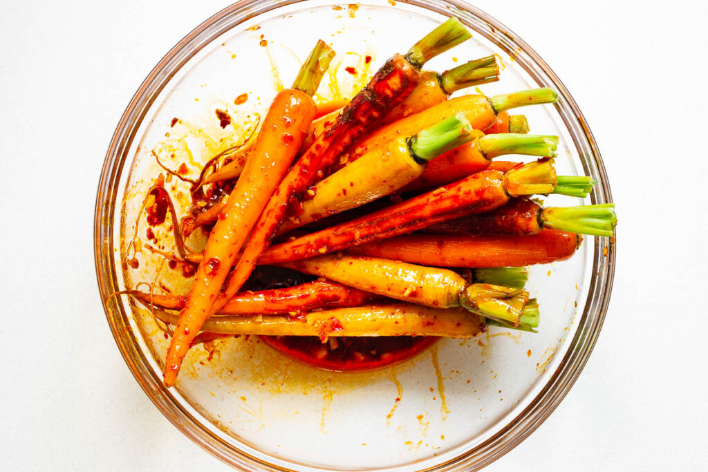Rainbow carrots tossed in harissa and maple marinade in a glass bowl.