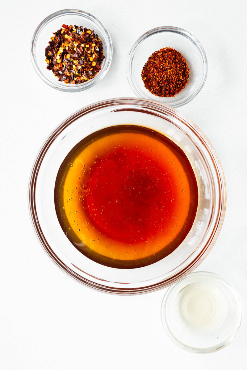 Hot honey ingredients viewed from above on a white surface, including chilli flakes, honey and apple cider vinegar.