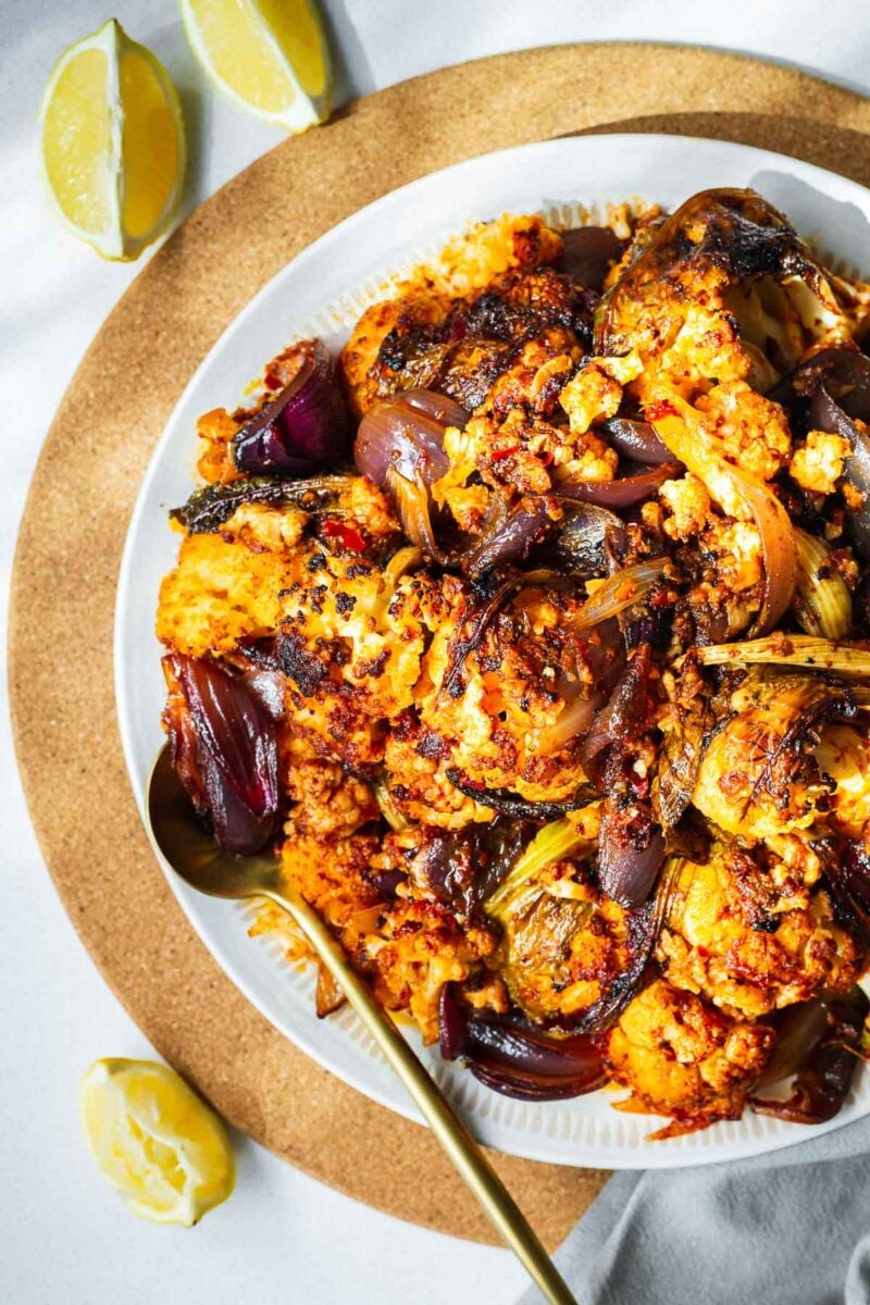 Top down view of a plate of harissa roasted cauliflower with a gold serving spoon and lemon wedges.