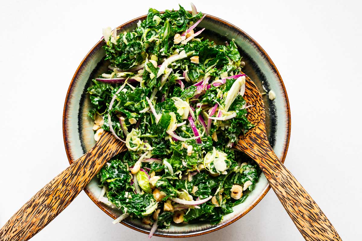 Tossing kale salad with a creamy tahini dressing using a pair of wooden salad tongs.