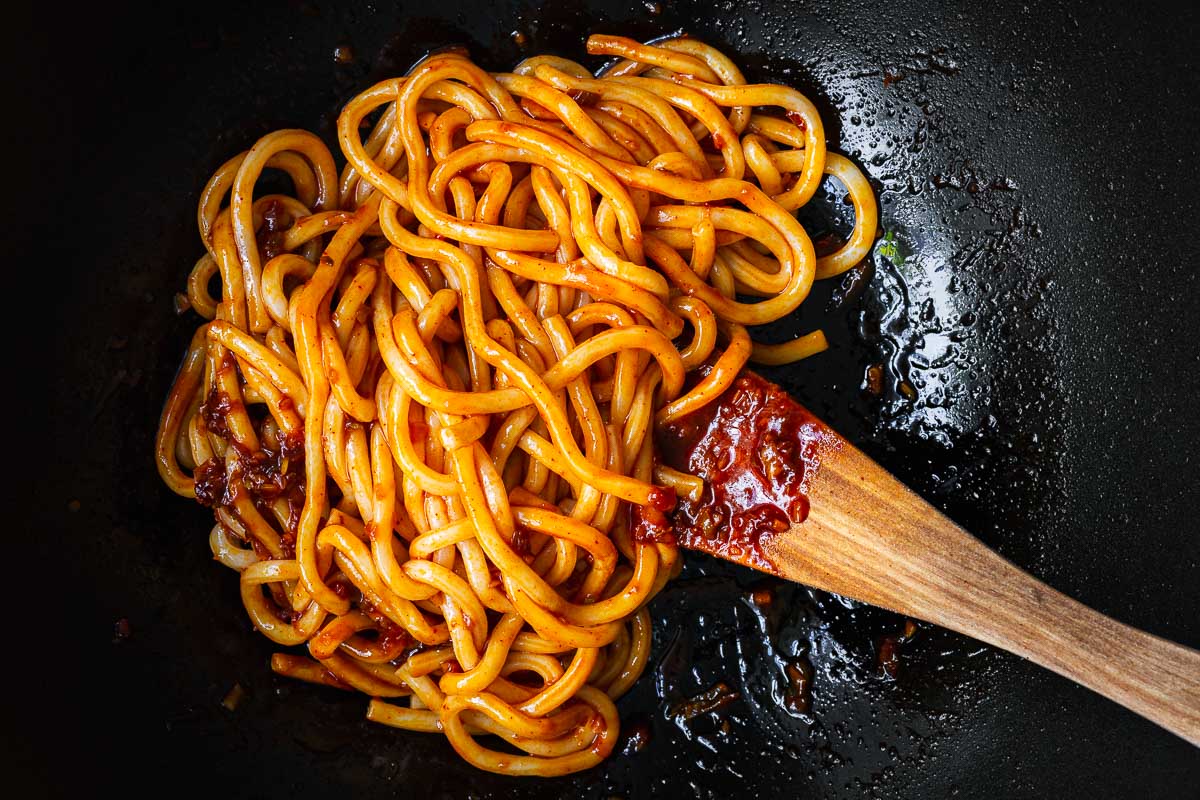Cooked udon noodles getting coated in gochujang noodle sauce in a wok.