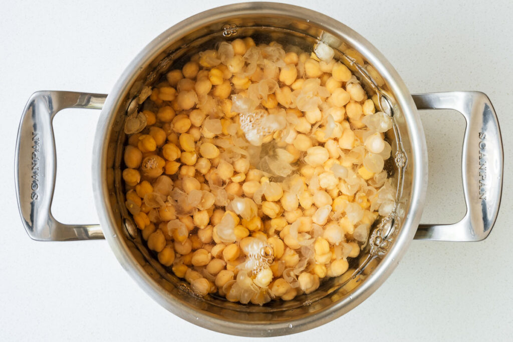 Chickpeas in a pot with water and chickpea skins floating to the top.
