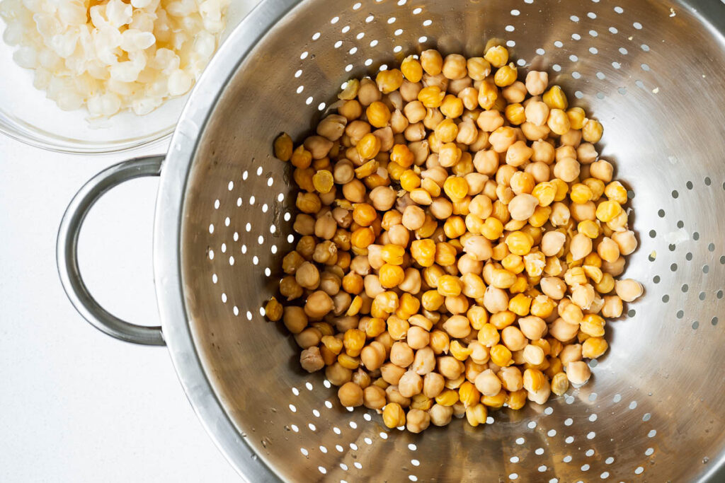 Cooked chickpeas draining in a colander next to a small bowl of chickpea skins.
