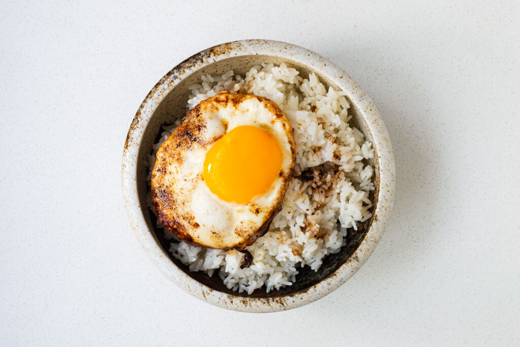 A ceramic bowl with cooked white rice topped with a fried egg with crunchy edges and runny yolk.