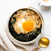 A ceramic bowl with fried rice, fried egg with a runny yolk, crushed seaweed and toasted sesame seeds, drizzled with soy sauce, sesame sauce and brown butter.