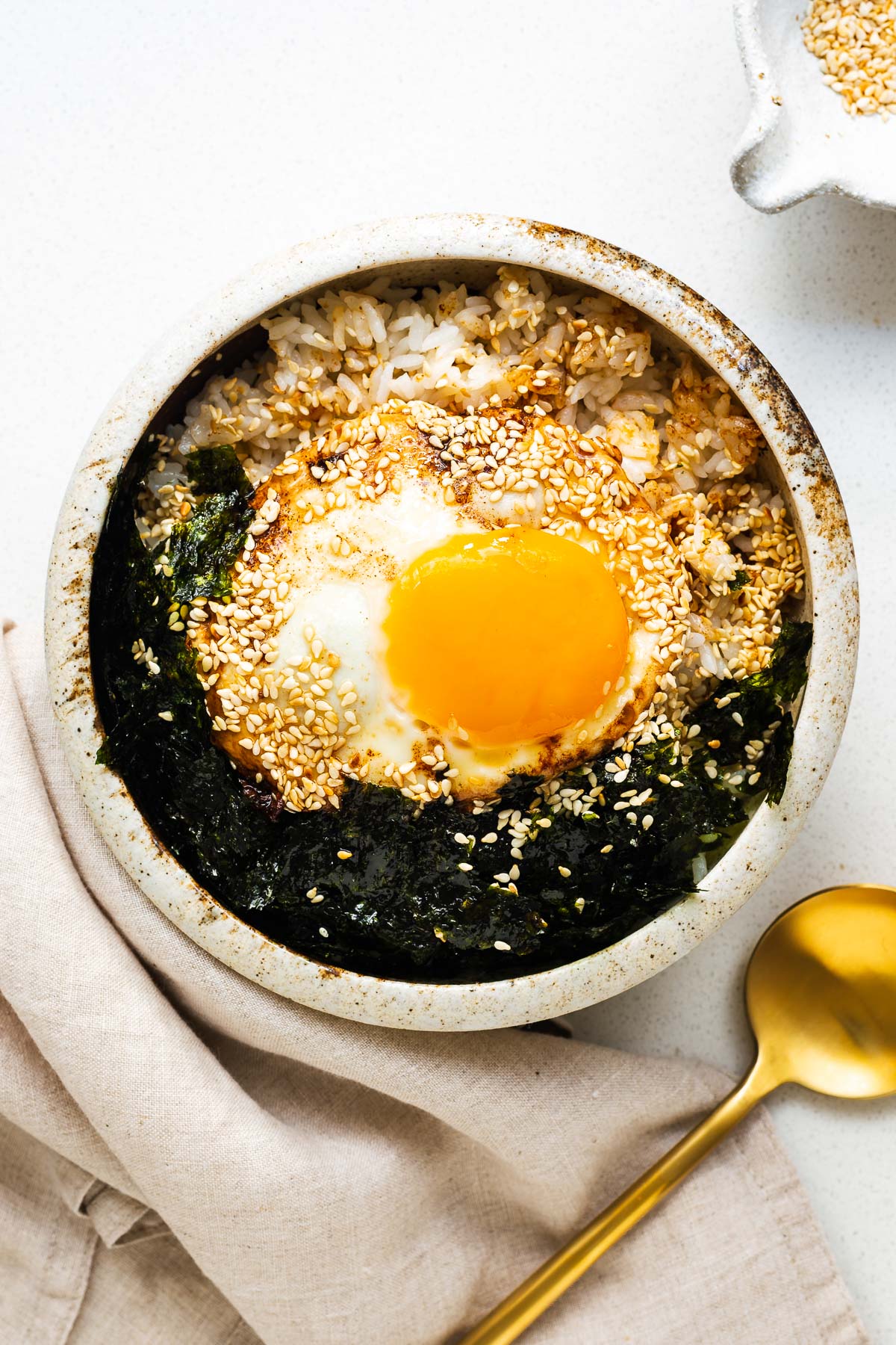 A soy sauce fried egg on rice with crushed gim (roasted seaweed) and sesame seeds in a ceramic bowl.