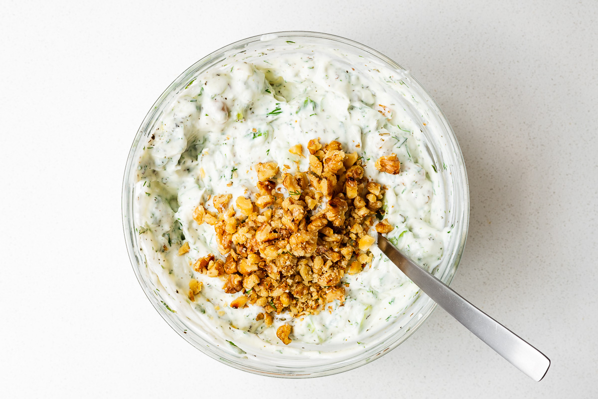 Mixing Persian cucumber and yoghurt dip with walnuts in a medium-sized glass bowl.