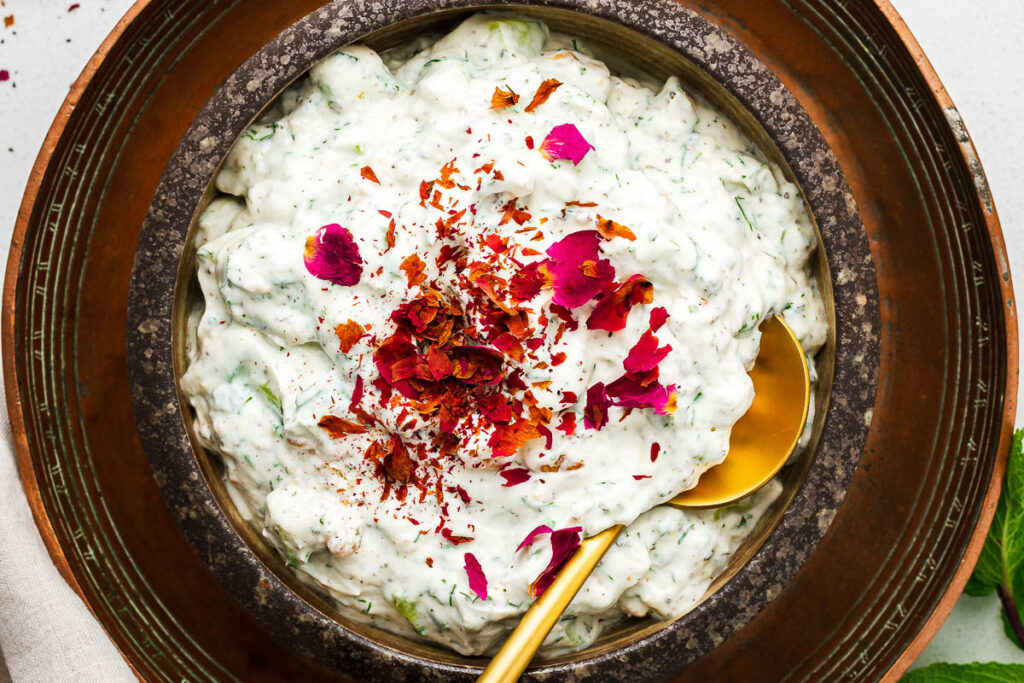 A horisontal close-up image of Persian yoghurt salad with cucumbers in a brown ceramic bowl, topped with pink dried rose petals.