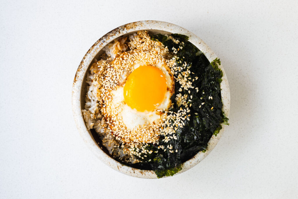 An assembled gyeran bap breakfast bowl with buttered rice, fried egg, roasted seaweed and toasted sesame seeds drizzled with soy sauce and sesame oil.