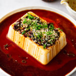 A block of silken tofu surrounded by a soy-based dressing with a soy sauce bowl and a bottle of chili crisp visible in the back ground.