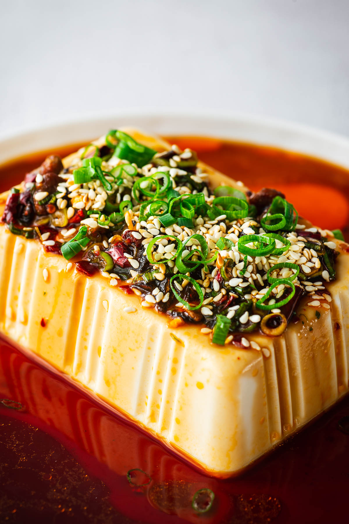 A close-up of silken tofu with a sweet and spicy soy dressing and toppings such sliced green onions, chili crisp and toasted sesame seeds.