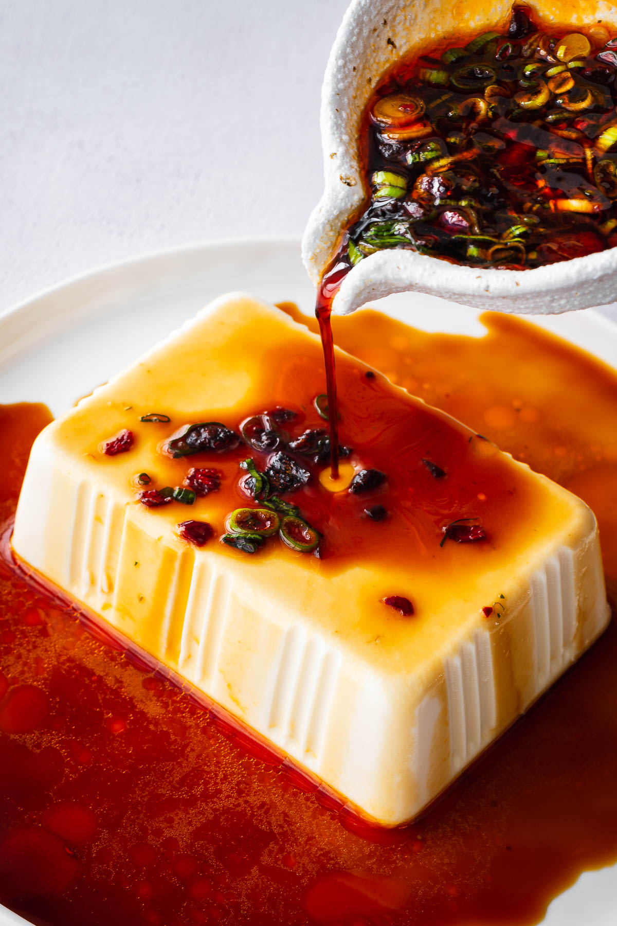 A block of silken tofu surrounded by a soy-based dressing being poured from a soy sauce bowl.