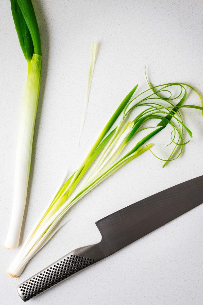 A green onion sliced in half and then into thing strips next to a chefs knife.