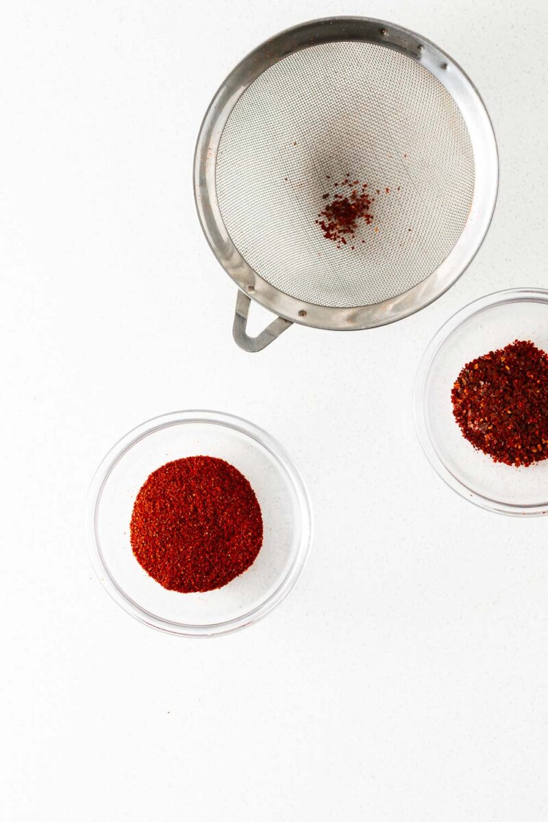 A small fine mesh strainer with two small bowls. One contains fine chilli powder, the other contains coarser chilli flakes.