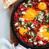 Harissa shakshuka with bright red roasted pepper sauce with soft-cooked eggs and visibly runny yolks topped with feta and parsley in a cast-iron skillet.