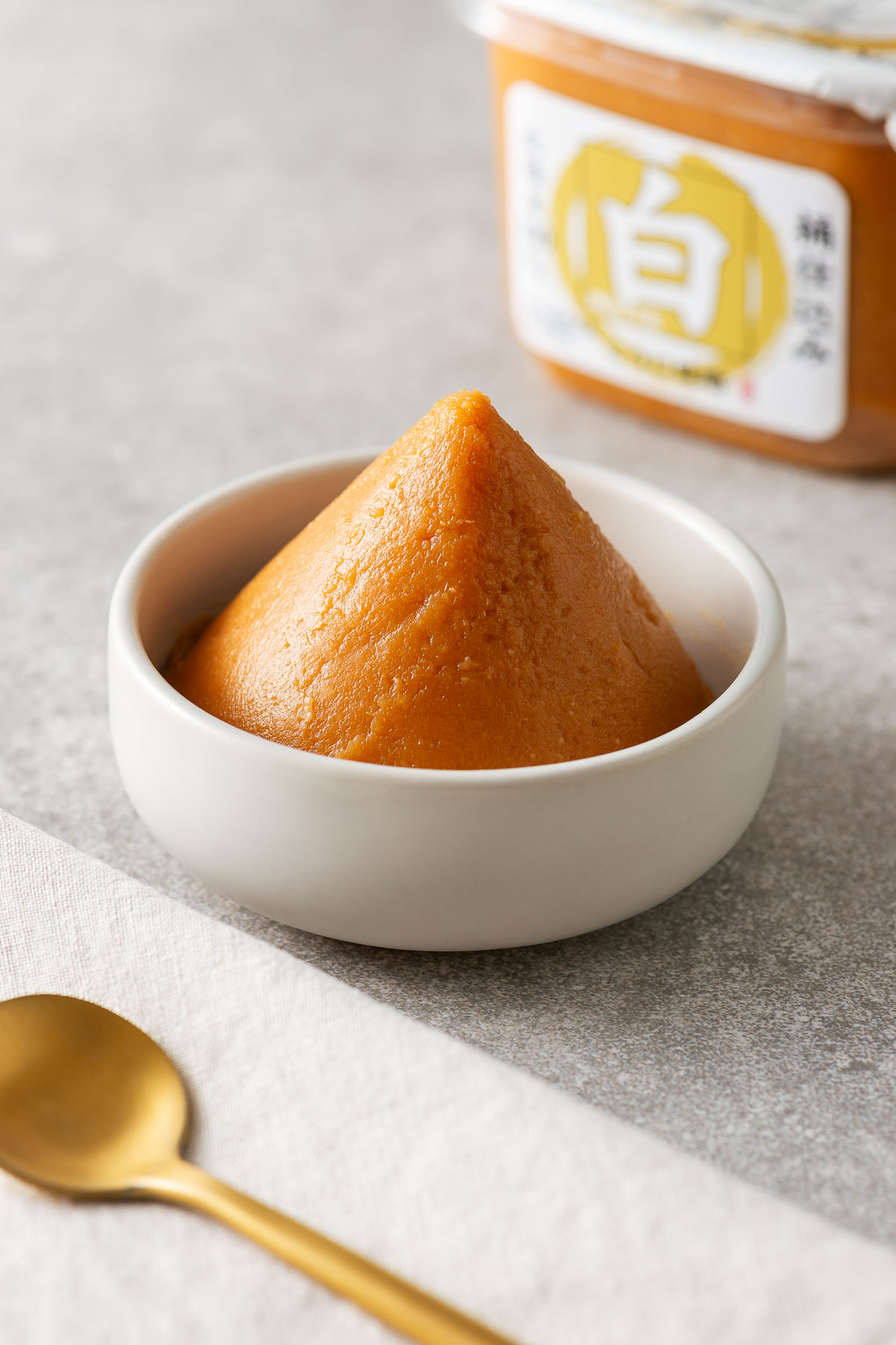 Whit miso paste shaped into a triangle in a small white bowl next to a tub of white miso paste.