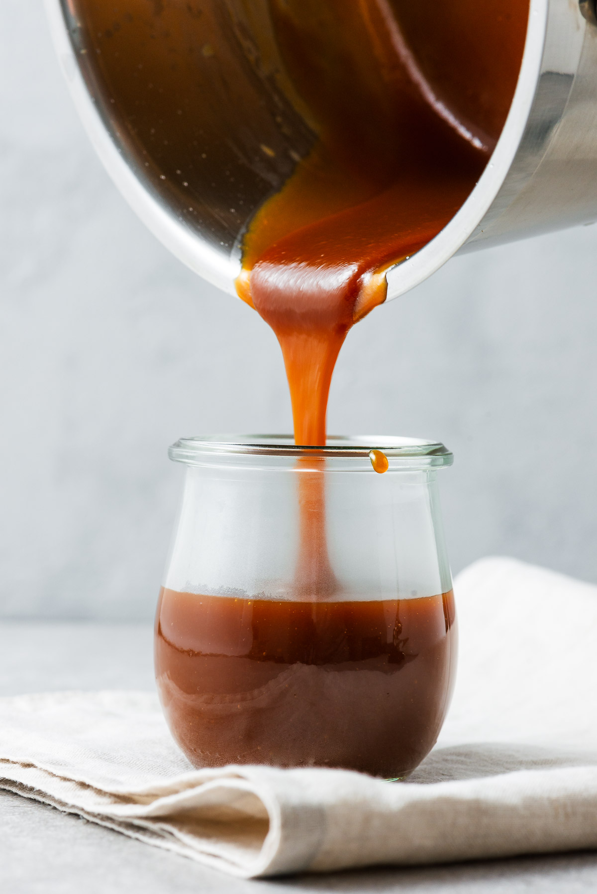 Pouring homemade miso caramel sauce from a stainless steel saucepan into a small glass jar.