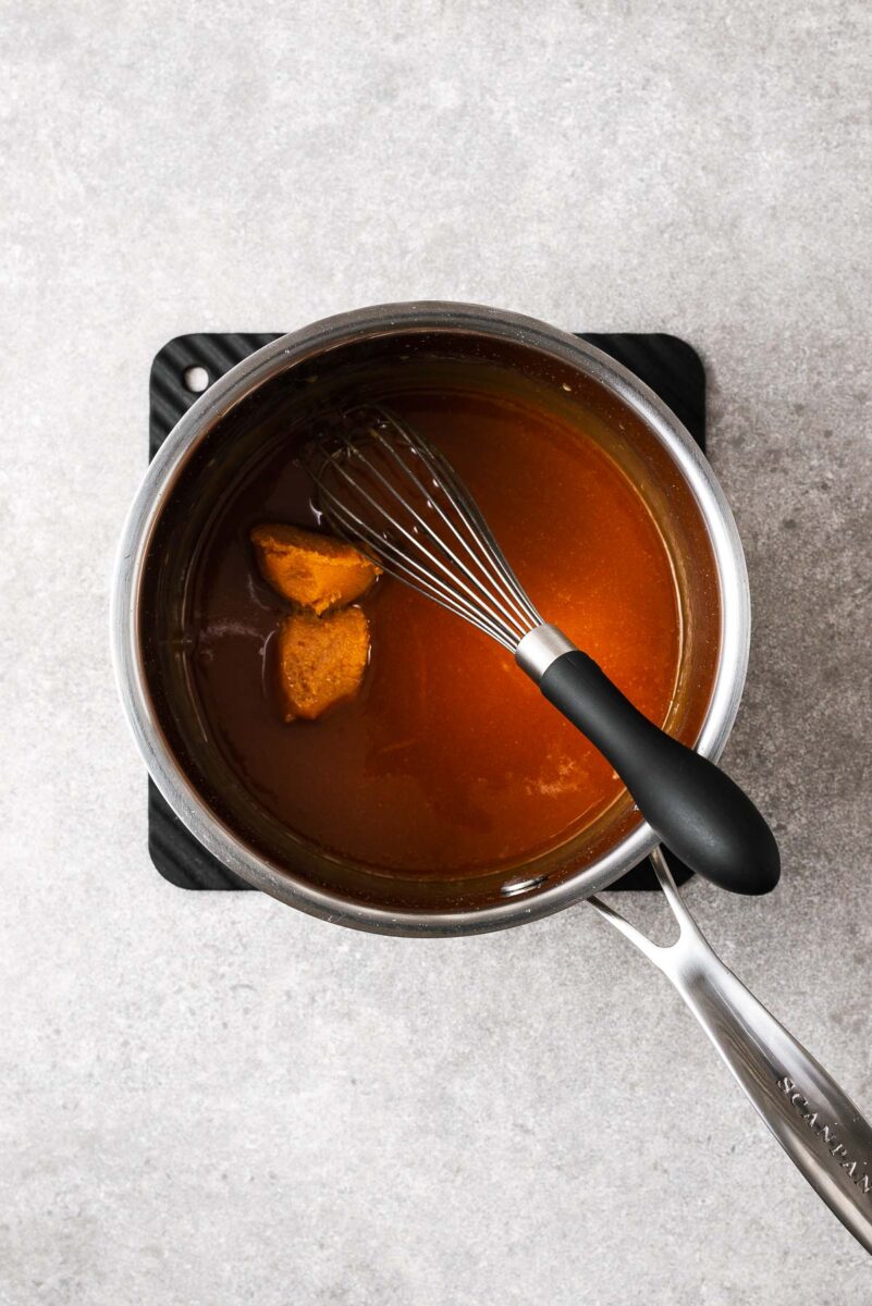 A stainless steel saucepan and balloon whisk with caramel and two tablespoons of white miso paste.