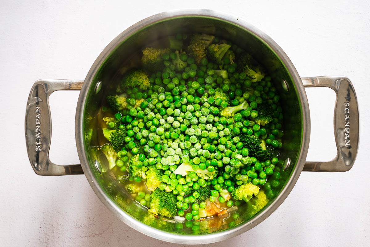 A stainless steel soup pot with broccoli, aromatics, vegetable broth and frozen peas.
