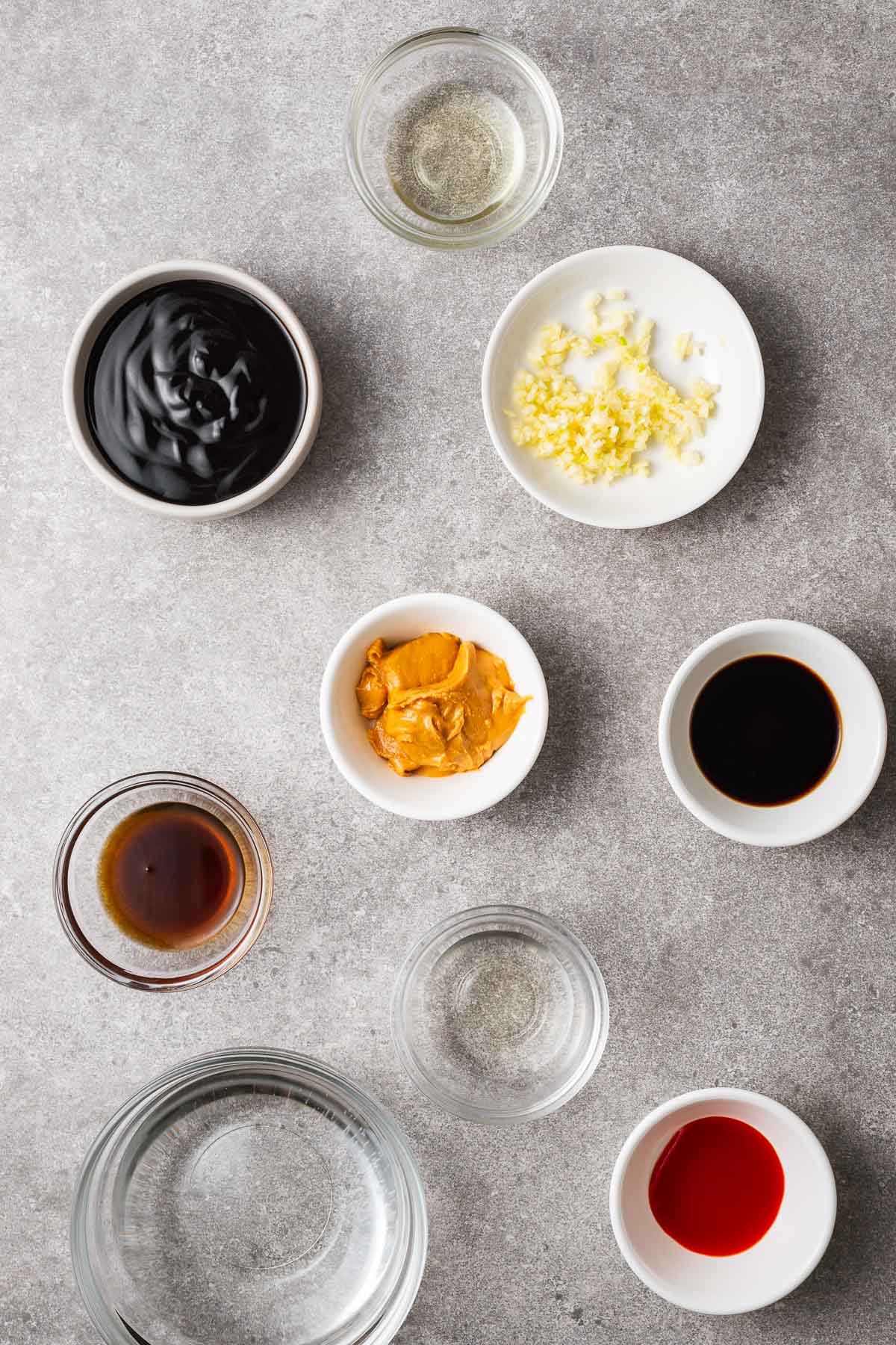 Ingredients for hoisin peanut sauce arranged in small bowls including hoisin sauce, peanut butter, minced garlic, toasted sesame oil, sriracha sauce, rice vinegar and water.