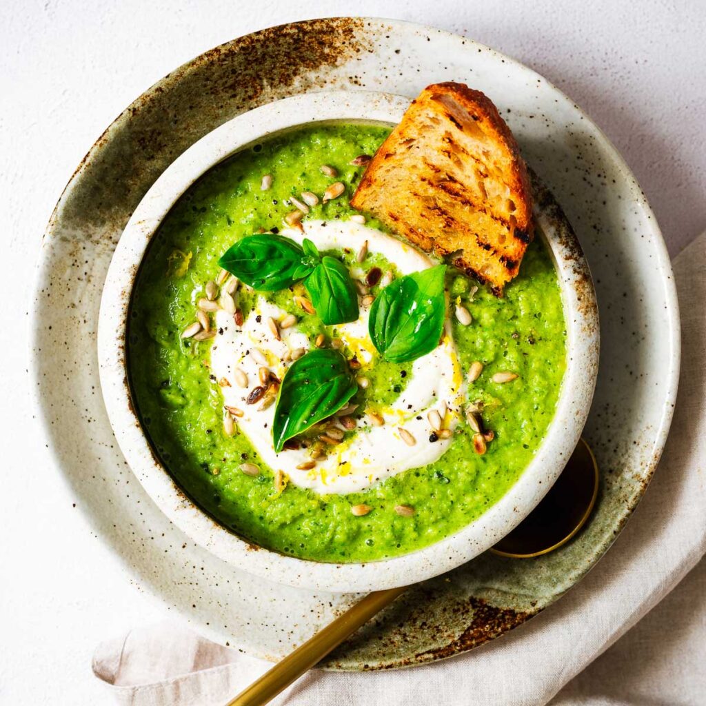 A bowl of pea and broccoli soup with fresh basil, toasted sunflower seeds and creamy cashew sauce with a slice of toasted sourdough bread.