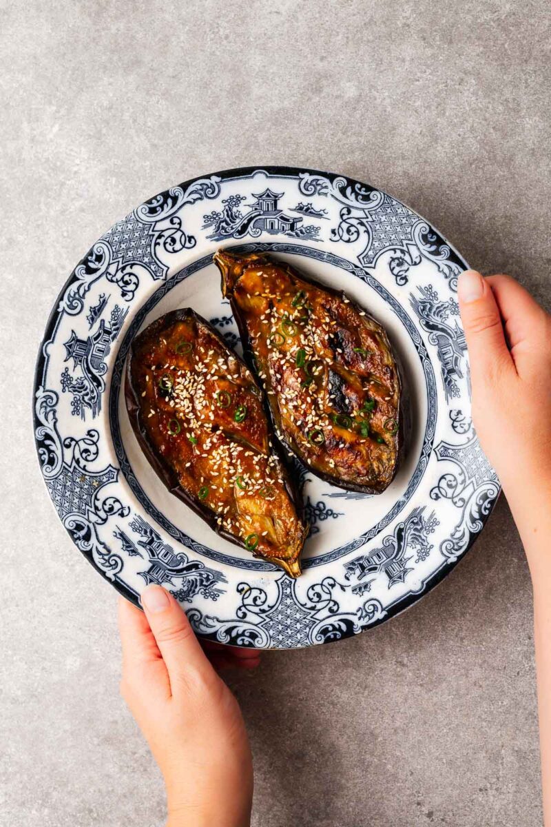 Miso eggplant (miso aubergine) sprinkled with sesame seeds and green onions served in a traditional Japanese ceramic bowl framed with a pair of hands.