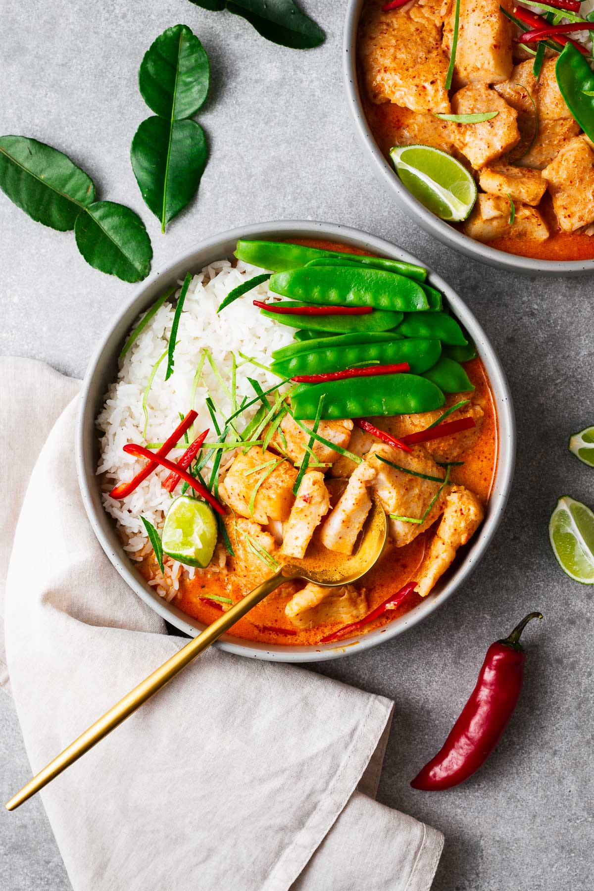 A bowl with steamed jasmine rice, Thai fish red curry and snow peas topped with sliced red chile and kaffir lime leaves wit a golden serving spoon.