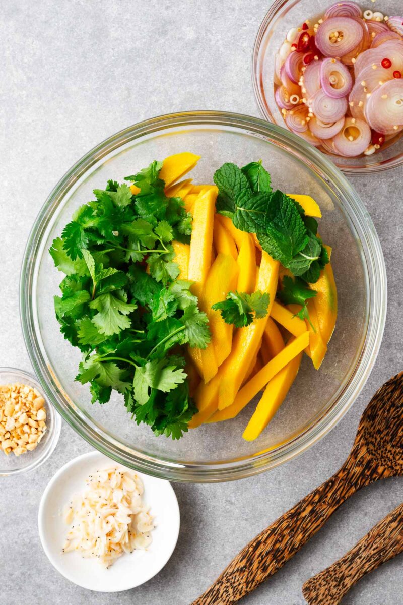 A large mixing bowl with thinly sliced green mango strips and fresh herbs.