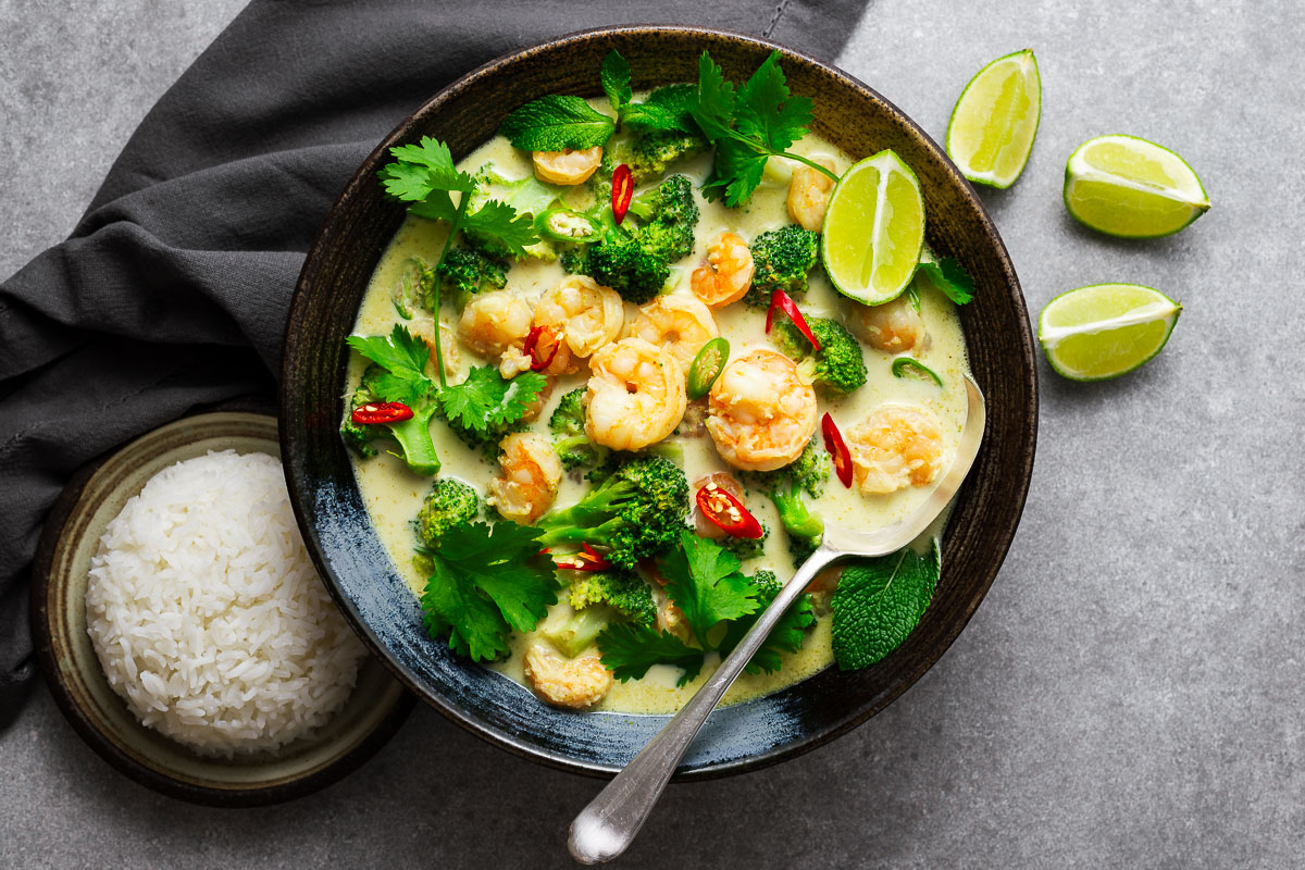 A Thai green curry bowl with shrimp and green vegetables garnished with fresh herbs and lime wedges with a serving spoon and a side or rice.