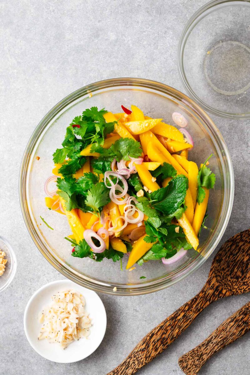 Assembling the Thai mango salad in a large glass bowl with the salad ingredients and salad dressing.