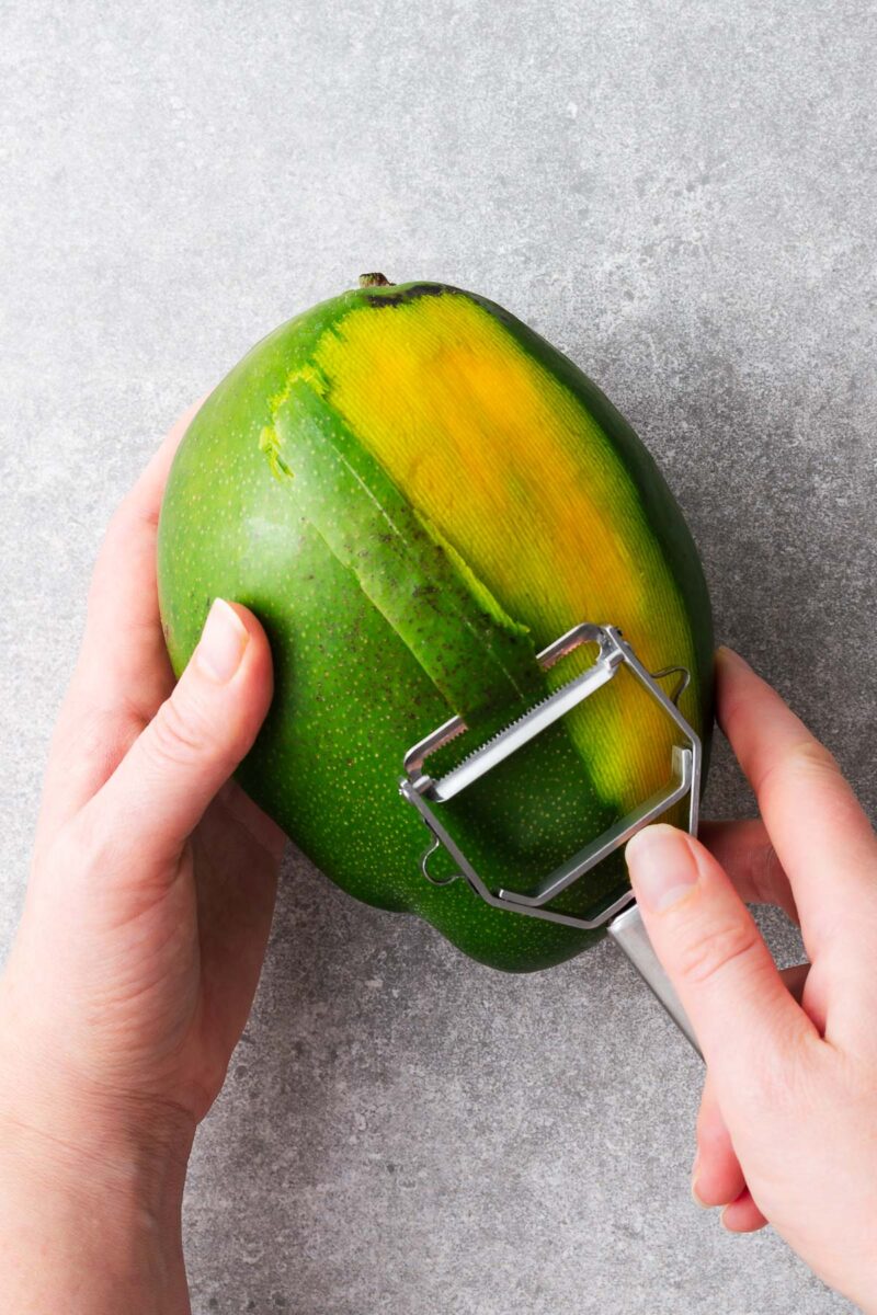 Two hands peeling a green mango with a vegetable peeler.