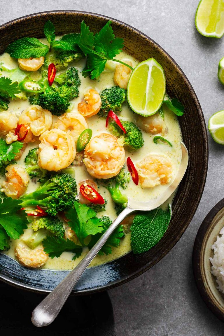 A ceramic bowl with Thai green curry with shrimp and broccoli garnished with fresh herbs and lime wedges with a serving spoon.