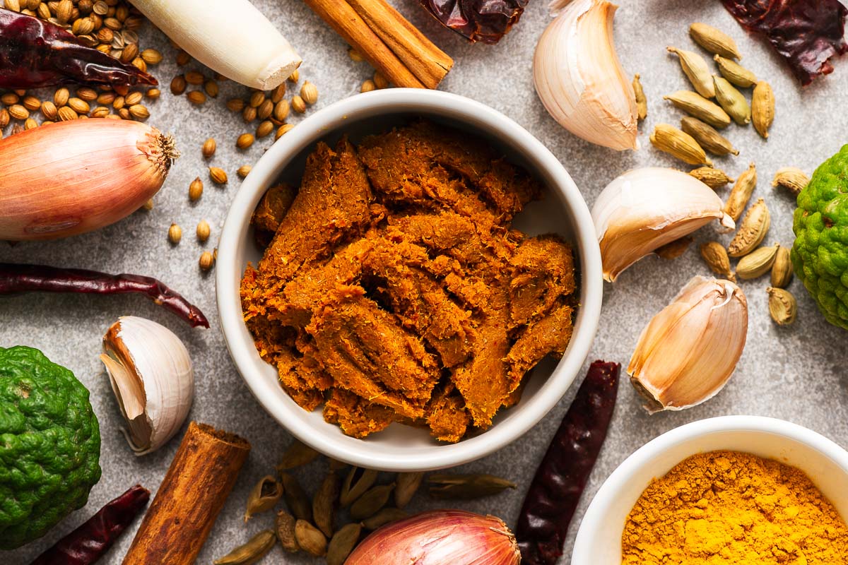 A bowl with Thai yellow curry paste surrounded by key ingredients like dried red chillies, kaffir lime, lemongrass, garlic, shallots, and spices like turmeric, curry powder, coriander seeds, and cinnamon.