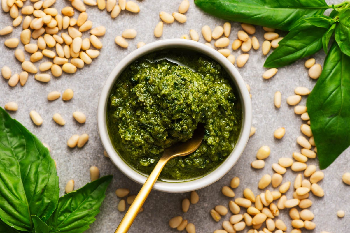 A small ceramic bowl with homemade Genoese pesto surrounded by pine nuts and fresh basil leaves.