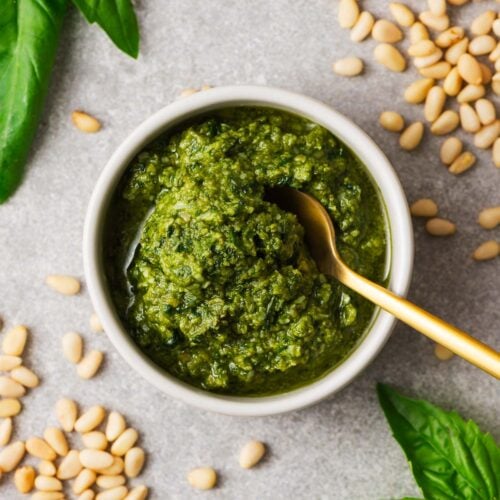 Traditional Italian basil pesto in a white bowl surrounded by typical pesto ingredients including pine nuts and Genovese basil.